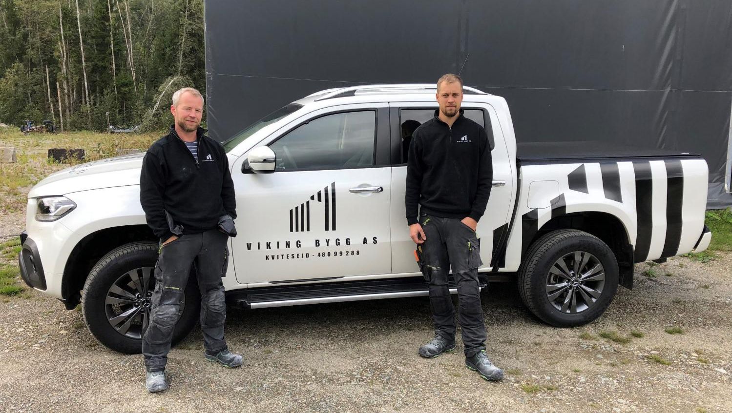 To arbeidere kledd i svart genser med firmalogo står foran svart og hvit firmabil med logo